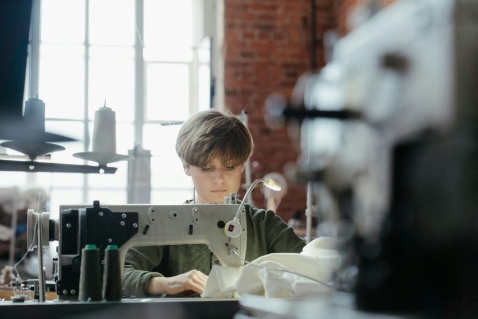 En person syr i vitt tyg med en industrisymaskin i en lokal med tegelväggar.