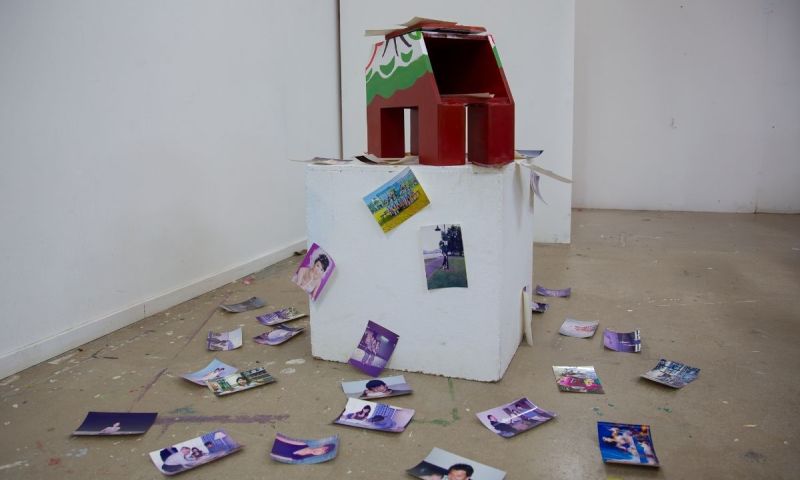 Installation i galleri. En trähäst på en vit låda omgiven av fotografier utspridda på golvet.