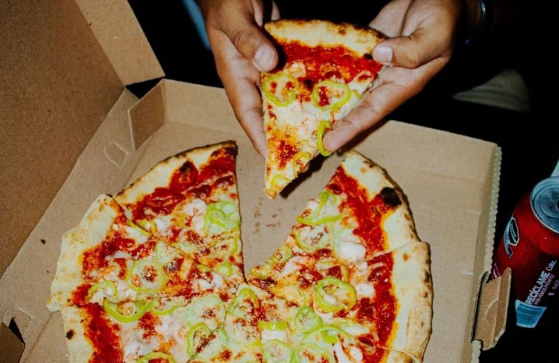 En hand lyfter en pizzaslice ur kartongen medan resten av pizzan ligger kvar