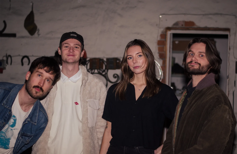 Fyra personer står uppställda och ser in i kameran i en enkel replokalsmiljö