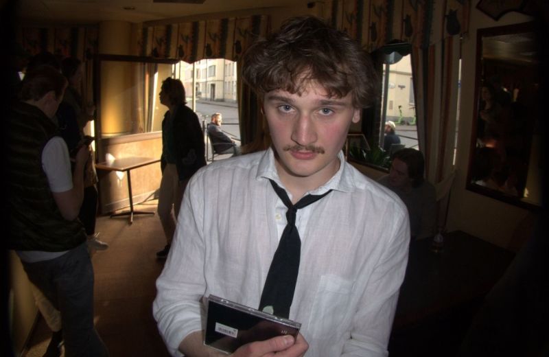 En närbild på poeten Albin Carlsson som håller ett CD-fodral, omgiven av människor i en lokal.