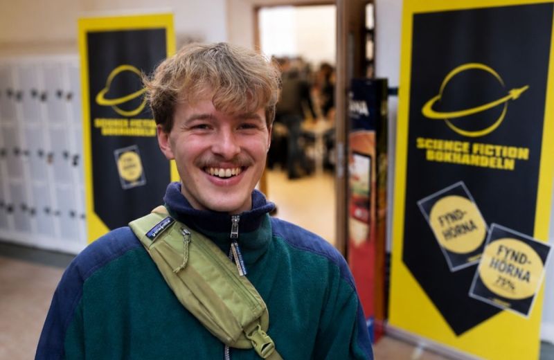 En leende person i grön fleece och axelväska står framför skyltar för Science Fiction Bokhandeln. Bakgrunden är lätt suddig med entré och besökare bakom.