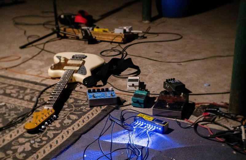 En vit elgitarr ligger på golvet omgiven av effektpedaler och sladdar i en replokal.