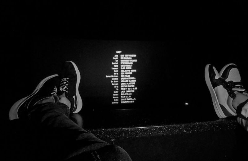 Svartvit bild på fötter som vilar mot en rad i en biosalong medan eftertexterna rullar på duken.