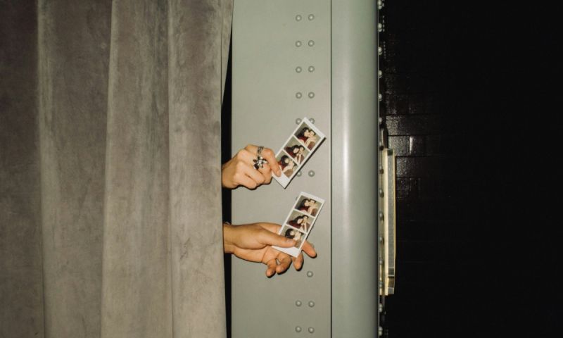 Händer som sticker ut bakom ett sammetsdraperi och håller upp två fotoremsor från en fotoautomat.
