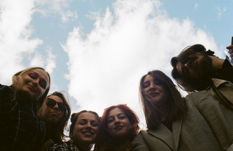 Sex ungdomar tittar ner i kameran underifrån mot en ljusblå himmel med vita moln.