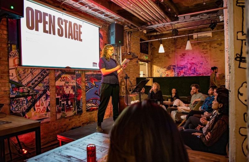 En person läser ur en bok på en liten scen framför en publik. En projektorskärm visar texten ”OPEN STAGE”.