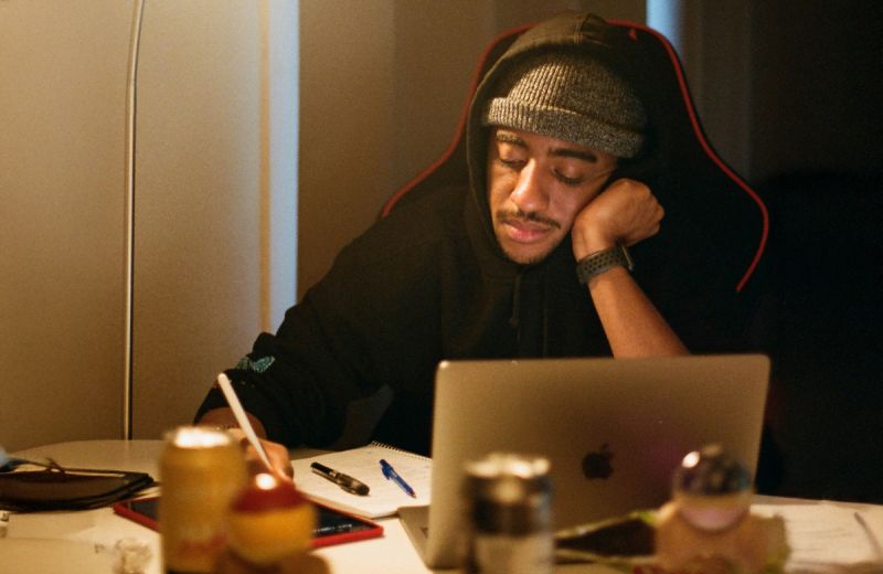 Person i hoodie och mössa skriver i ett block framför en bärbar dator.