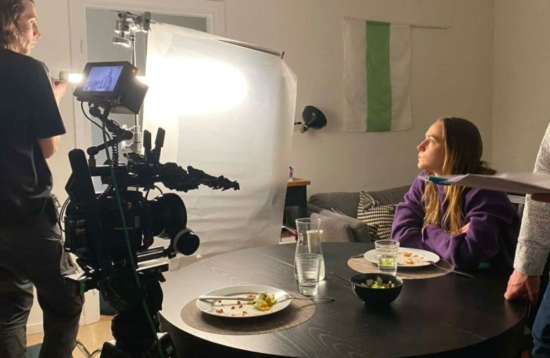 Koncentrerad kvinna i lila tröja vid ett matbord filmas med stor kamera och lampa