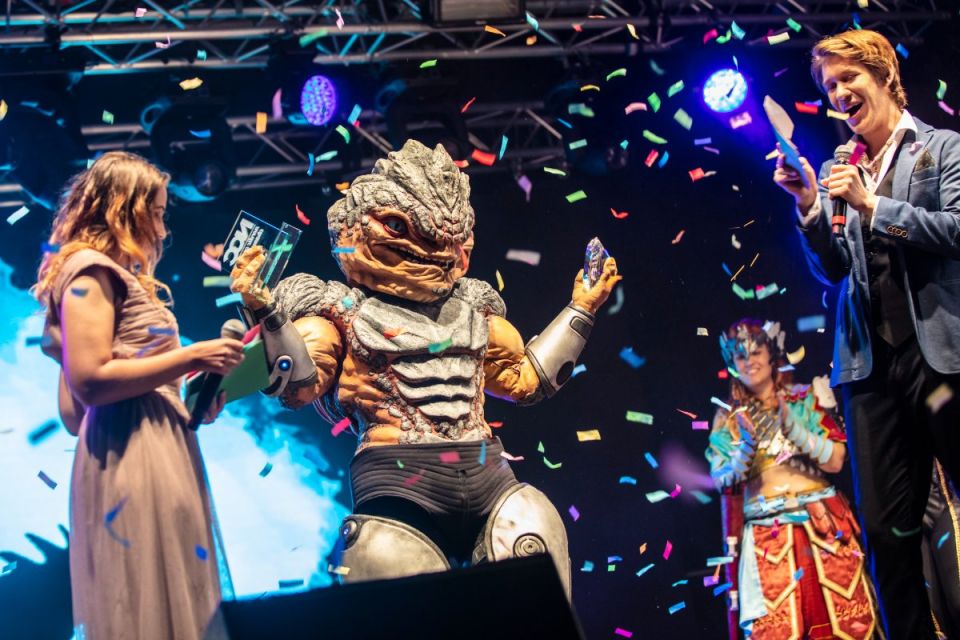 En cosplayer i detaljrik reptilliknande rustning håller upp två priser på en scen medan färgglad konfetti regnar ner och personer runt omkring firar.