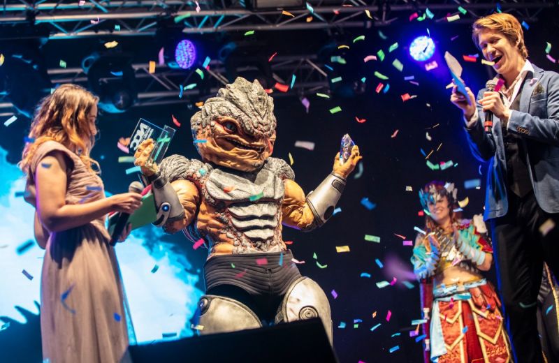 En cosplayer i detaljrik reptilliknande rustning håller upp två priser på en scen medan färgglad konfetti regnar ner och personer runt omkring firar.