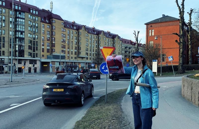Ung tjej i blå jacka står vid en trafikerad väg och visar stolt upp en affisch med text.