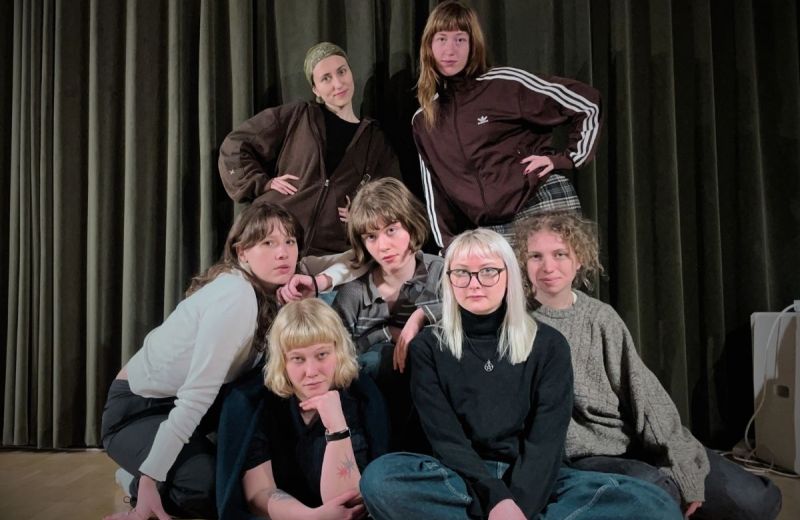 Nära gruppbild på sju unga vuxna i olika stilar, mot mörk drakplagg.