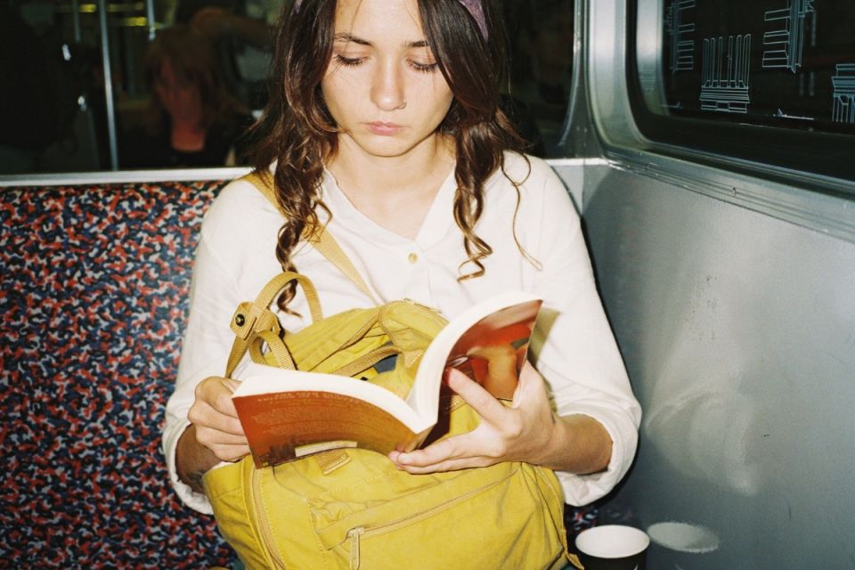 En person sitter på en tunnelbana och läser en bok med en gul ryggsäck i knäet.